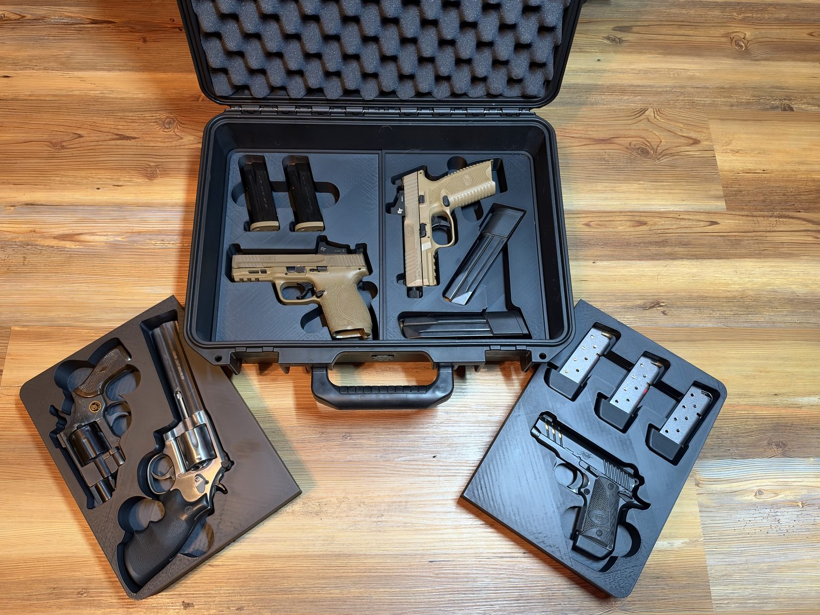 An open hard gun case with a two-pocket printed insert holding two tan pistols and magazines, plus two loose printed trays: one with revolvers, one with a compact pistol and magazines.